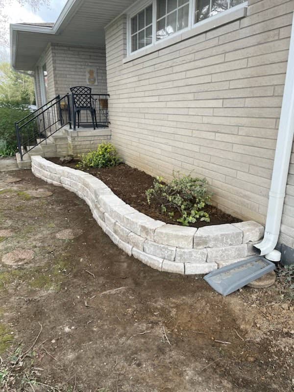 Curved raised garden bed with light stone retaining wall along the foundation of a brick house, filled with fresh soil and small green shrubs.