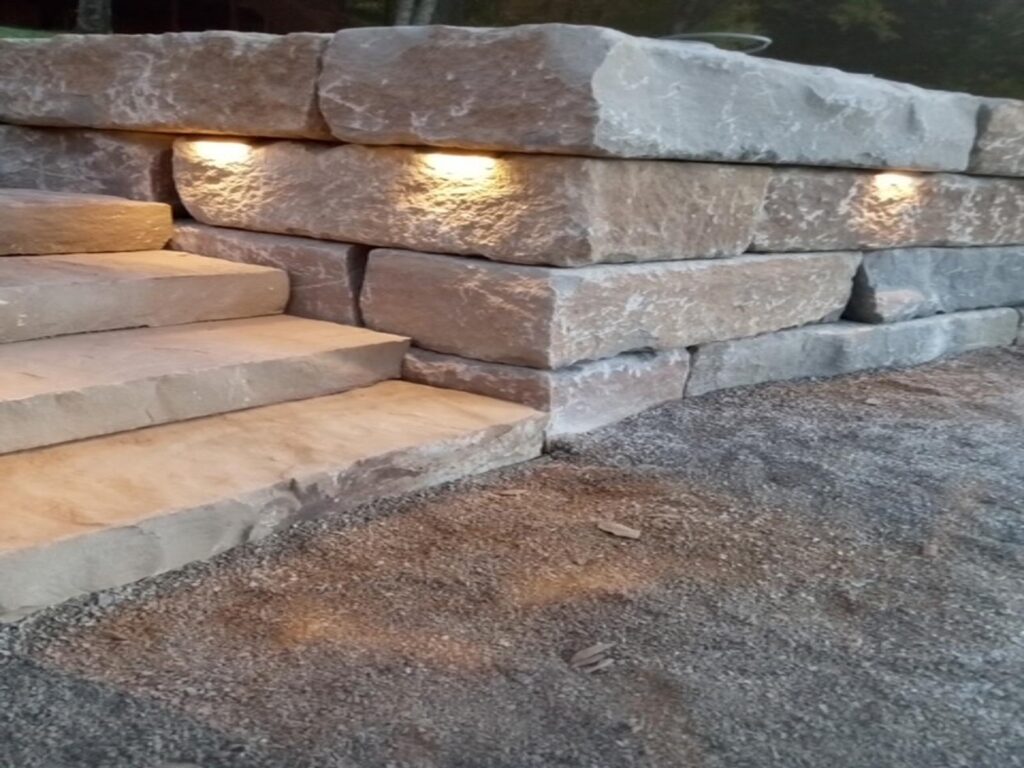 A close-up of a rustic stone retaining wall with built-in warm-toned lighting, alongside large stone steps leading upward, set against a gravel ground surface.