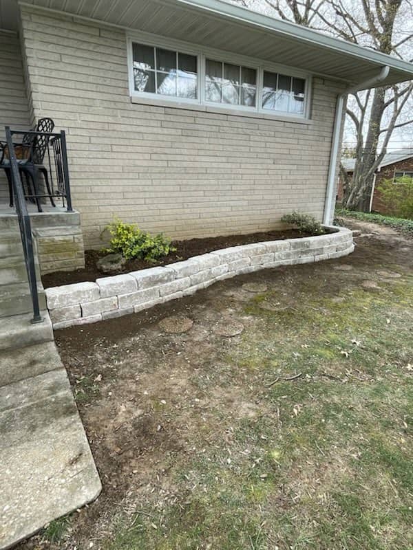Newly built raised garden bed with light stone retaining wall along the side of a brick house, featuring a few small shrubs and fresh soil.
